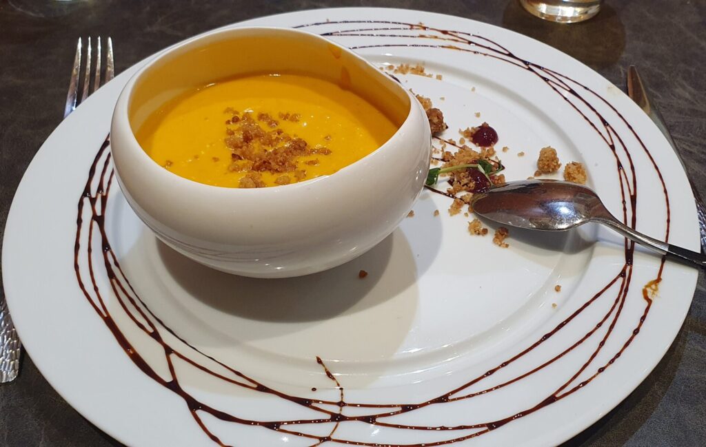 En Français: "veloute de potimarron et sa crumble á la châtaigne" In English: "Butternut squash soup with chestnut crumble" A bowl of thick and creamy pumpkin soup, topped with a crumble of roasted caramelised chestnuts and a balsamic drizzle on the plate.