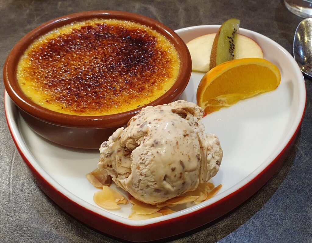 En Français: "Notre emblématique créme brulée et sa boule au glace de Daim" In English: "Our iconic crème brûlée and its Daim bar ice cream" A dish of thick and creamy custard with its signature toasted sugary top, this brulee calls to you to dive in. Off to the side, a scoop of Daim bar ice cream, that is both sweet and soothing with its crunchy Daim bar pieces.