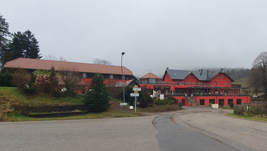 View of Hôtel le Velleda du Donon, on the edge of Mont Donon, in the Bas-Rhin of France. Picture shows the hotel from the front, the long and winding road sweeps up past the front of the hotel, with the majestic rose pink walls of the building rising into the sky, the steel grey slate roof looking dramatic against the pale grey sky.